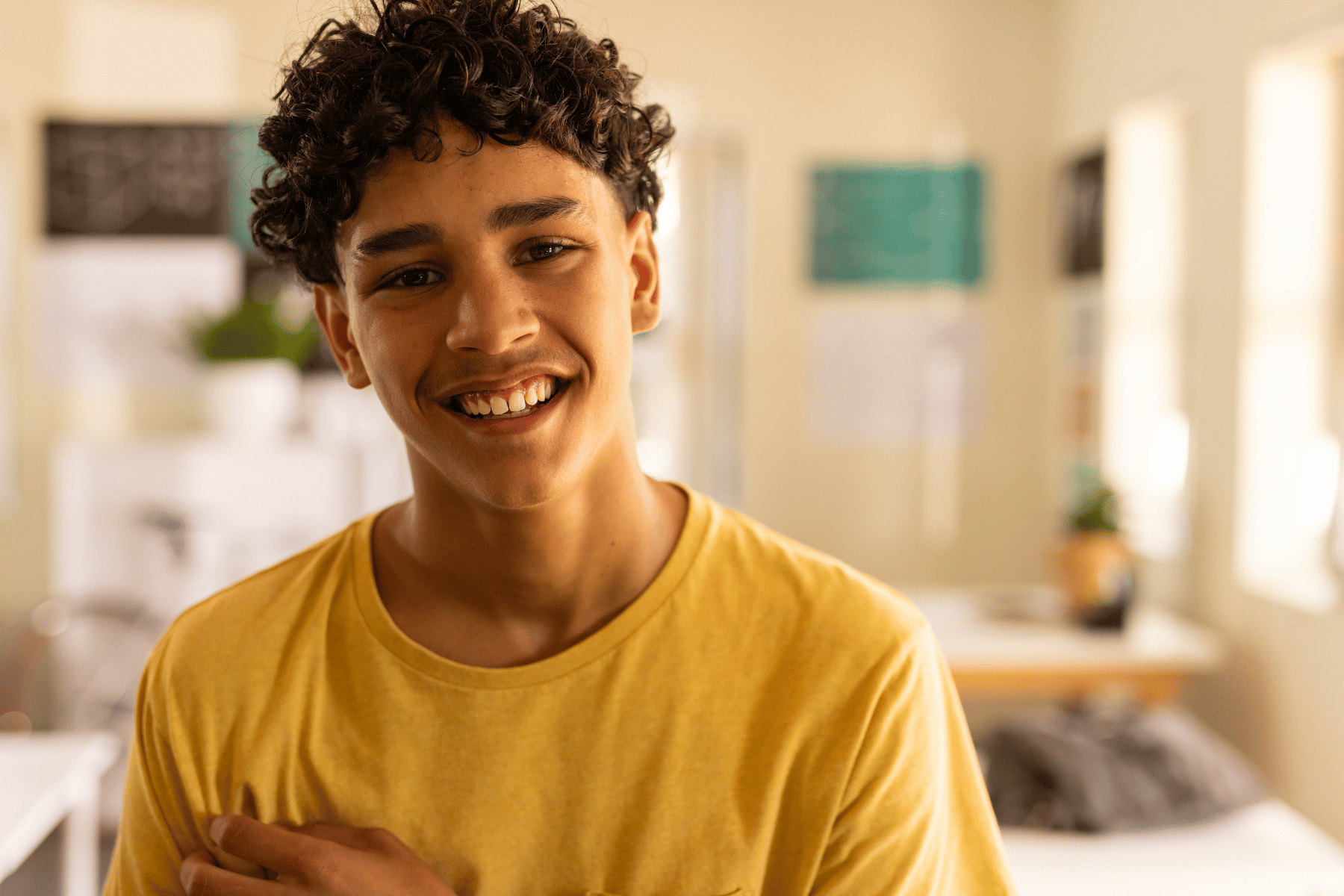 Smiling teenage boy with curly hair wearing a yellow shirt, standing in a warmly lit room with soft natural light and a cozy, home-like background.